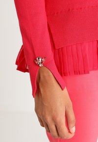 Manche d'un pull rose vif avec une fente au poignet et trois petites pierres précieuses roses au-dessus d'une couche de tissu plissé. Main détendue à côté.