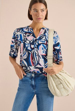 Vrouw die een blouse met abstract patroon en blauwe spijkerbroek draagt, met een grote crèmekleurige schoudertas tegen een effen beige achtergrond.
