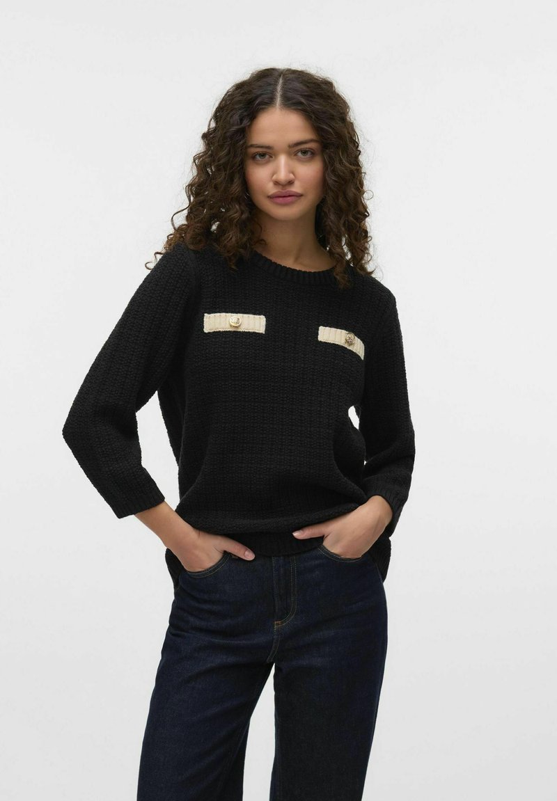 Zwarte gebreide trui met driekwart mouwen, met twee crèmekleurige zakaccenten en gouden knopen, gecombineerd met donkerdenim jeans.