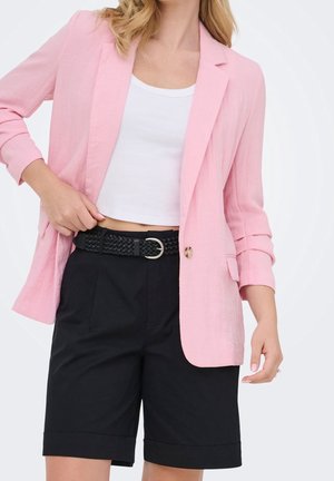 Femme portant un blazer rose, un débardeur court blanc, une ceinture tressée noire et un short noir longueur genou avec des revers roulés.