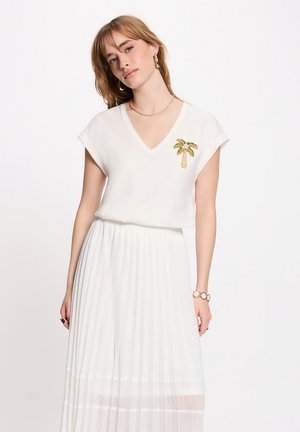 Donna che indossa una maglietta bianca a scollo a V con una toppa a palme e una gonna plissettata bianca, accessoriata con orecchini a cerchio dorati e un braccialetto.