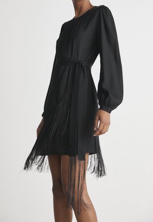 Femme portant une robe noire à manches longues avec une ceinture à la taille et un détail de frange le long de la jupe sur un fond uni.