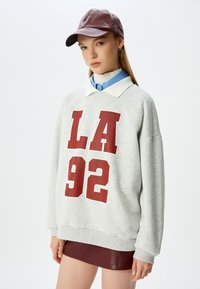 Sudadera gris con estampado rojo "LA 92", con un cuello blanco. Combinada con una falda de cuero burdeos y una gorra a juego.