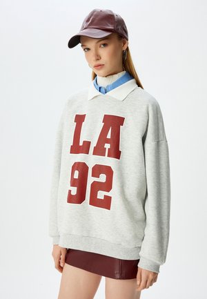 Sudadera gris con estampado rojo "LA 92", con un cuello blanco. Combinada con una falda de cuero burdeos y una gorra a juego.