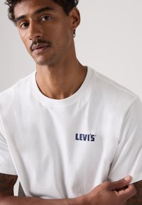Λευκό βαμβακερό μπλουζάκι με στρογγυλή λαιμόκοψη και μπλε λογότυπο Levi's στο στήθος. Το μοντέλο έχει τα χέρια σταυρωμένα, δείχνοντας τα κοντά μανίκια και την ομαλή υφή.
