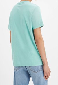 Osoba oblečená v světle modré košili s krátkým rukávem a světle modrých džínech, zobrazená zezadu na bílém pozadí.