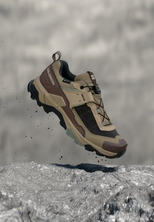 Scarpa da trekking Salomon marrone e nera con suola spessa che fluttua sopra un terreno roccioso con particelle di polvere in un ambiente naturale all’aperto.