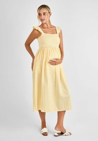 Vestido de maternidad de cuadros amarillos con cuerpo smock, escote cuadrado y tirantes con volantes. Falda fluida hasta el tobillo, combinada con sandalias planas.