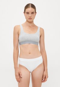 Woman with slicked-back hair wearing a grey sports bra and white high-waisted underwear, standing against a plain white background.