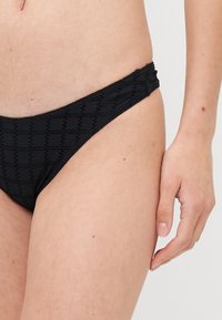 Crne badića s teksturiranim, kockastim uzorkom. Ima detalje s nabranim stranama i nisko strukiranje za minimalnu pokrivenost.