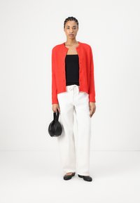 Red knitted cardigan with buttons, worn over a black top. Paired with white trousers and a black handbag. Flat black shoes complete the look.