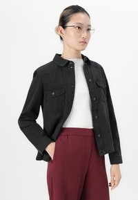 Young woman wearing glasses, black buttoned jacket over white top, and high-waisted burgundy pants, standing with one hand in pocket.