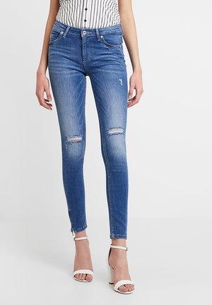 Jeans ajustados de mezclilla azul con detalles desgastados, dos desgastes en las rodillas, una textura suave y un ajuste ceñido. Los dobladillos con cremallera acentúan el diseño.