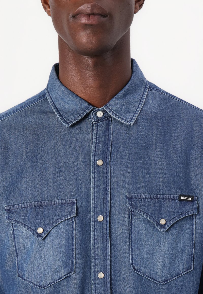 Hombre vestido con una camisa de mezclilla azul, de botones y con cuello puntiagudo, que tiene dos bolsillos en el pecho con botones a presión, uno de los cuales está etiquetado "Replay".