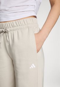 Gaiši bēšas sporta bikses, izgatavotas no mīksta auduma, ar elastīgu jostasvietu un auklu. Vīles pie kabatas rotā balts Adidas logo.