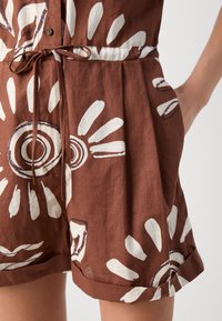 Rompers in lino marrone con motivi circolari e a foglia bianchi e marroni scuri, dotato di vita regolabile con coulisse e risvolti per uno stile alla moda.
