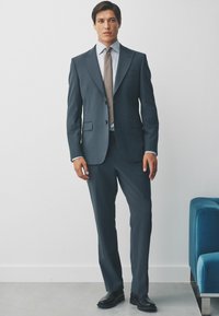 Dunkelblauer Anzug mit zwei Knöpfen, Revers und seitlichen Taschen. Weißes Hemd und gemusterte Krawatte. Glänzende schwarze Schnürschuhe.