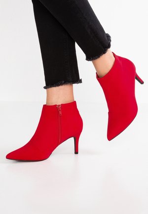 Czerwone szpiczaste botki na wysokim obcasie sięgające kostki, zapinane na boczne zamki, noszone z czarnymi obcisłymi dżinsami z postrzępionym wykończeniem u dołu.