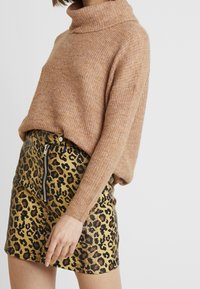 Lichtbruine, gebreide trui met een hoge col, gecombineerd met een minirok met luipaardprint, voorzien van een zwarte rits en gouden accenten.