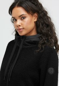 Black fleece jacket with a textured finish, featuring a zip-up neck, adjustable black drawstrings, and a round logo patch on the sleeve.