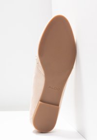 Escarpin en suede beige avec un bout arrondi et une semelle en caoutchouc brun légèrement texturée. Présente de petites clous dorés près du talon.