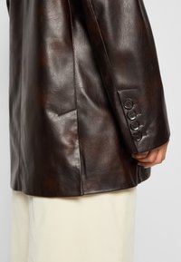 Blazer din piele maro cu un croi atent, textură fină și patru butoane decorative la manșetă. Asortat cu o țesătură de culoare crem dedesubt.