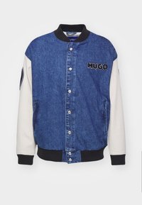 Kurtka dżinsowa z niebieskim korpusem, białymi rękawami i czarnymi prążkowanymi mankietami. Posiada zatrzaski i haftowane logo "HUGO" na piersi.
