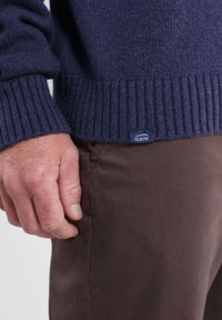 Pull en tricot marine avec des poignets et un ourlet côtelés, arborant une étiquette Oxbow. Un pantalon en coton marron est partiellement visible. Texture lisse.