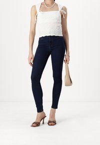 Vit smockad crop top med knytband, mörkblå högmidjade skinny jeans, bruna klackiga mules med nitar och en beige axelväska.