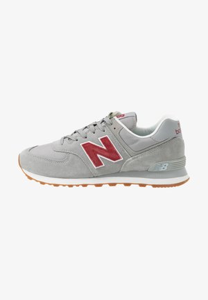 Szary i czerwony sneaker New Balance z cholewką zamszową i siateczkową, białą podeszwą środkową i gumową podeszwą zewnętrzną, widziany od zewnętrznej strony.