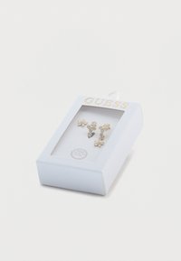 Σκουλαρίκια με φυτικό μοτίβο σε χρυσό τόνο με διαφανείς πολύτιμους λίθους, τοποθετημένα σε μια λευκή υφασμάτινη θήκη με το λογότυπο "GUESS" στο επάνω μέρος.