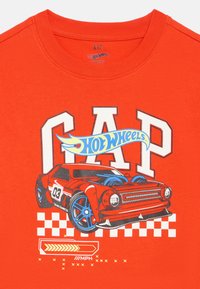 Červené detské tričko s logami "GAP" a "Hot Wheels", obsahujúce detailné červené pretekárske auto s modrými kolesami a dizajnom kockovanej vlajky.