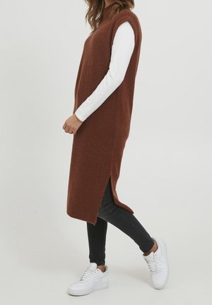 Femme marchant, portant une longue veste en tricot marron sans manches sur un haut blanc à manches longues, un jean noir et des baskets blanches sur un fond uni.