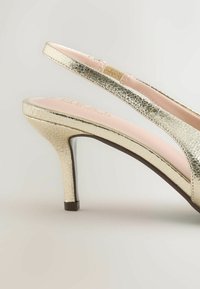 Chaussure à talon haut dorée brillante à bride arrière, avec une finition texturée, un talon fin et une semelle intérieure rembourrée rose pâle sur un fond neutre.