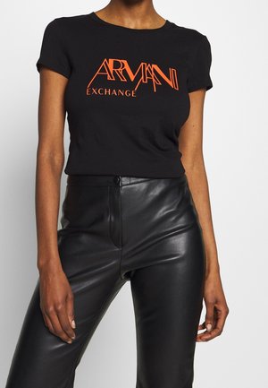 Femme portant un t-shirt noir à manches courtes avec un texte orange "Armani Exchange" et un pantalon en cuir noir taille haute.