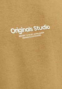 Barna pamut póló, fehér nyomtatott „Originals Studio” felirattal, alatta további szöveggel. Sima tapintás, egyszerű dizájn.