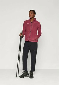 Sieviete burgundijas krāsas zip-up jakā un melnās biksēs, tur melnās pārgājienu nūjas, valkājot melnus pārgājienu zābakus, stāv pret vienkāršu fonu.