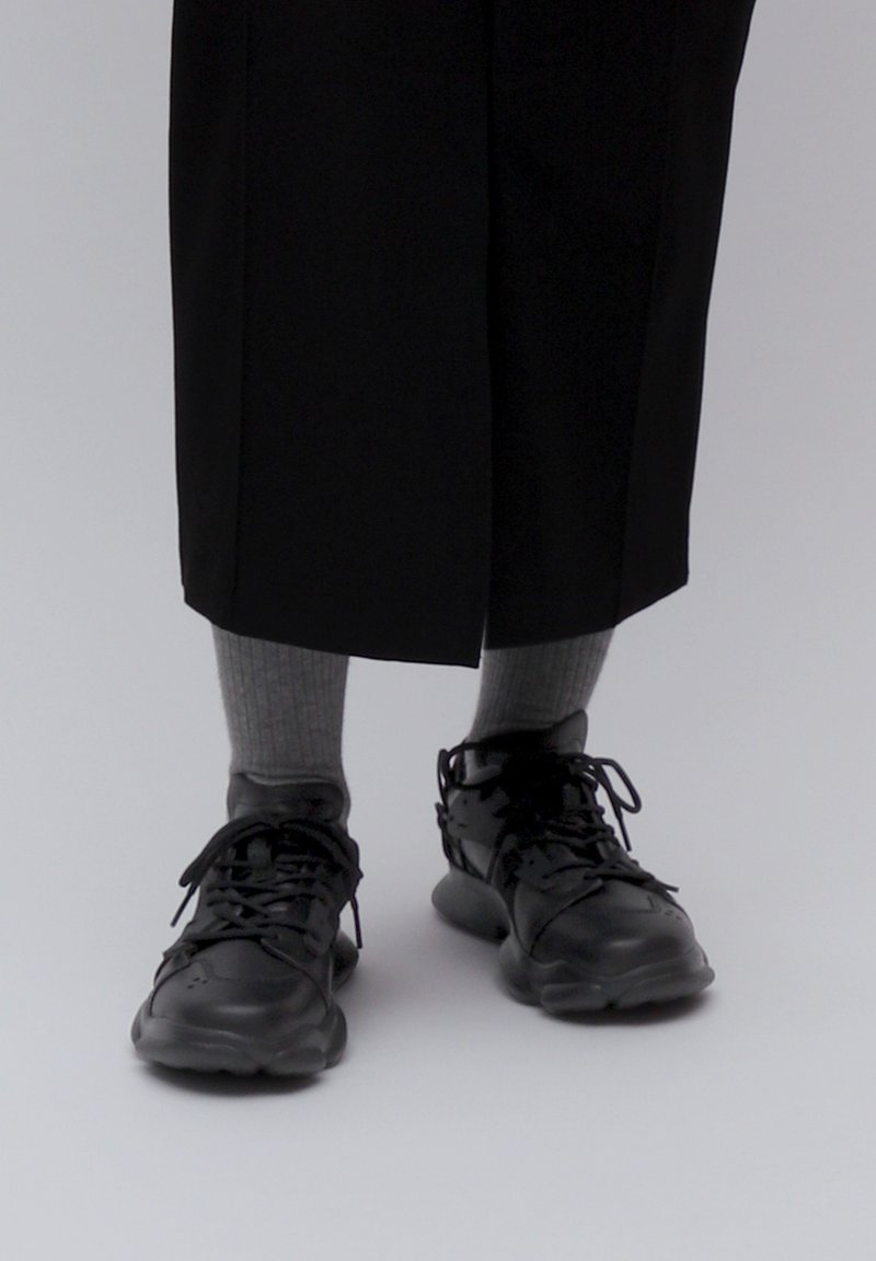 Black athletic shoes with textured soles, laces, and ventilation holes, paired with light gray knee-high socks, worn with black wide-leg pants.