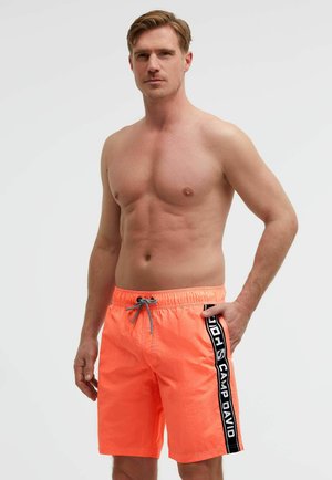 Shirtless man with short hair wearing orange swim shorts with black vertical stripe and white text on the side, standing against a plain background.