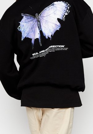 Sweat-shirt noir avec un grand papillon violet graphique et un texte blanc indiquant "Soul and Resurrection", porté avec un pantalon beige.
