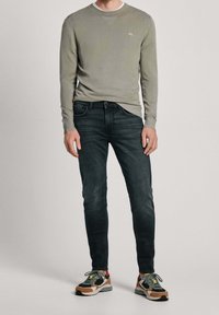 Ljusgrå långärmad skjorta med ribbad halsringning och subtil logotyp, i kombination med mörka slim-fit jeans och flerfärgade sneakers.