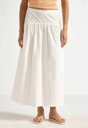 Kvinde iført en hvid tekstureret midinederdel, ferskenfarvet cropped top og beige flip-flops, stående mod en ensfarvet baggrund.