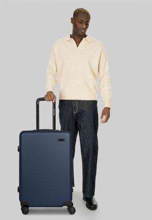 Hombre con suéter claro y jeans oscuros sujetando el asa de una maleta azul marino de cuatro ruedas sobre un fondo gris liso.