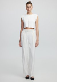 Witte crop top met een rits aan de voorkant, gecombineerd met witte high-waisted broek met een gestrikte riem. Zwarte platte schoenen maken de outfit af.