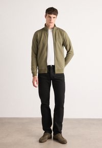 Giacca verde oliva con zip e colletto alto, indossata sopra una t-shirt bianca, abbinata a jeans neri e sneaker verdi olive su uno sfondo neutro.