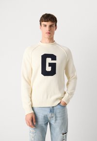 Cream knit sweater with a round neckline, ribbed cuffs, and hem, featuring a large navy "G" on the front. Worn with distressed blue jeans.