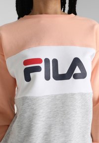 Persoon die een sweater draagt met kleurvlakken in roze, wit en grijs, met het FILA-logo in zwart en rood op de borst gedrukt.