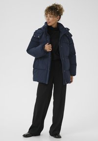 Moteris su garbanotais plaukais, vilkinti tamsiai mėlyną pūkinį švarką virš juodos turtleneko ir plačių juodų kelnių, stovi prieš neutralų foną.