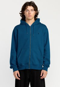 Sudadera con capucha y cremallera en color teal, que cuenta con un bolsillo canguro, puños y dobladillo acanalados, y un pequeño logo negro en el pecho.