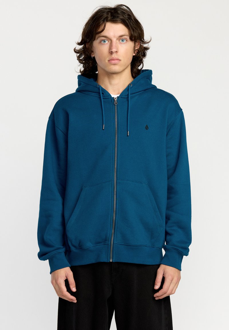 Sudadera con capucha y cremallera en color teal, que cuenta con un bolsillo canguro, puños y dobladillo acanalados, y un pequeño logo negro en el pecho.